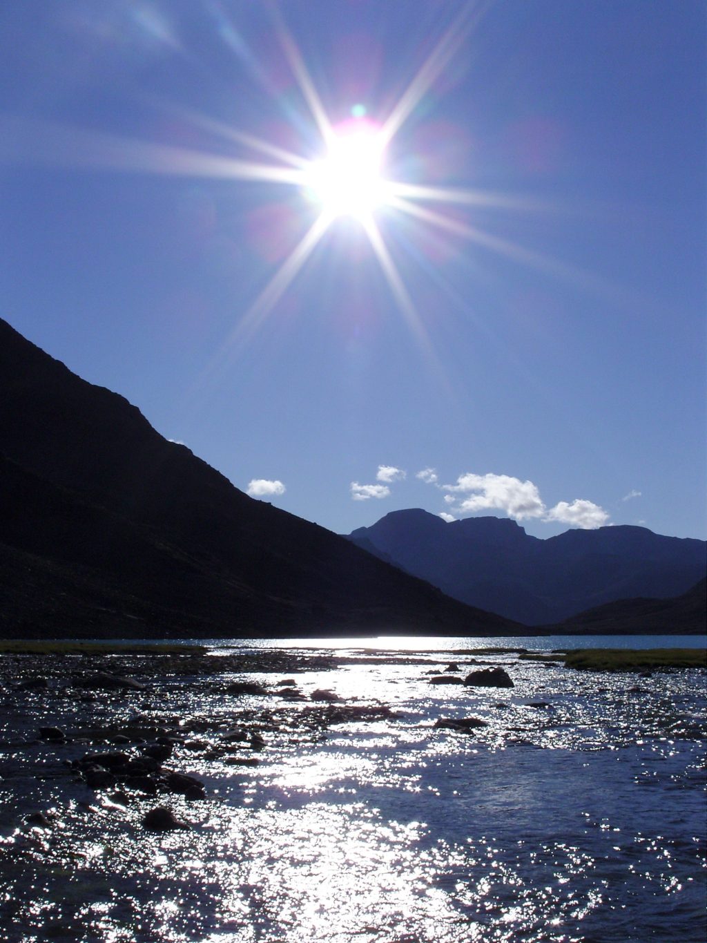 A body of water with mountains in the background and the sun shining brightly