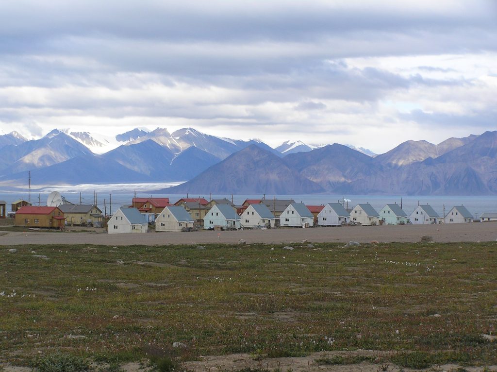 grass in the foreground with buildings further back infront of the sea. Mountains can be seen in the background, some with snow on them.