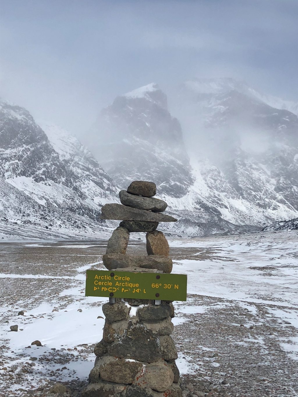 An inukshuk stone stack locating the Arctic Circle, with snow and mist covered mountains behind